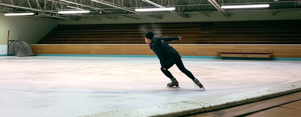 Lone figure skater on a small-town arena at morning practice, wooden bleachers and a folded net behind
